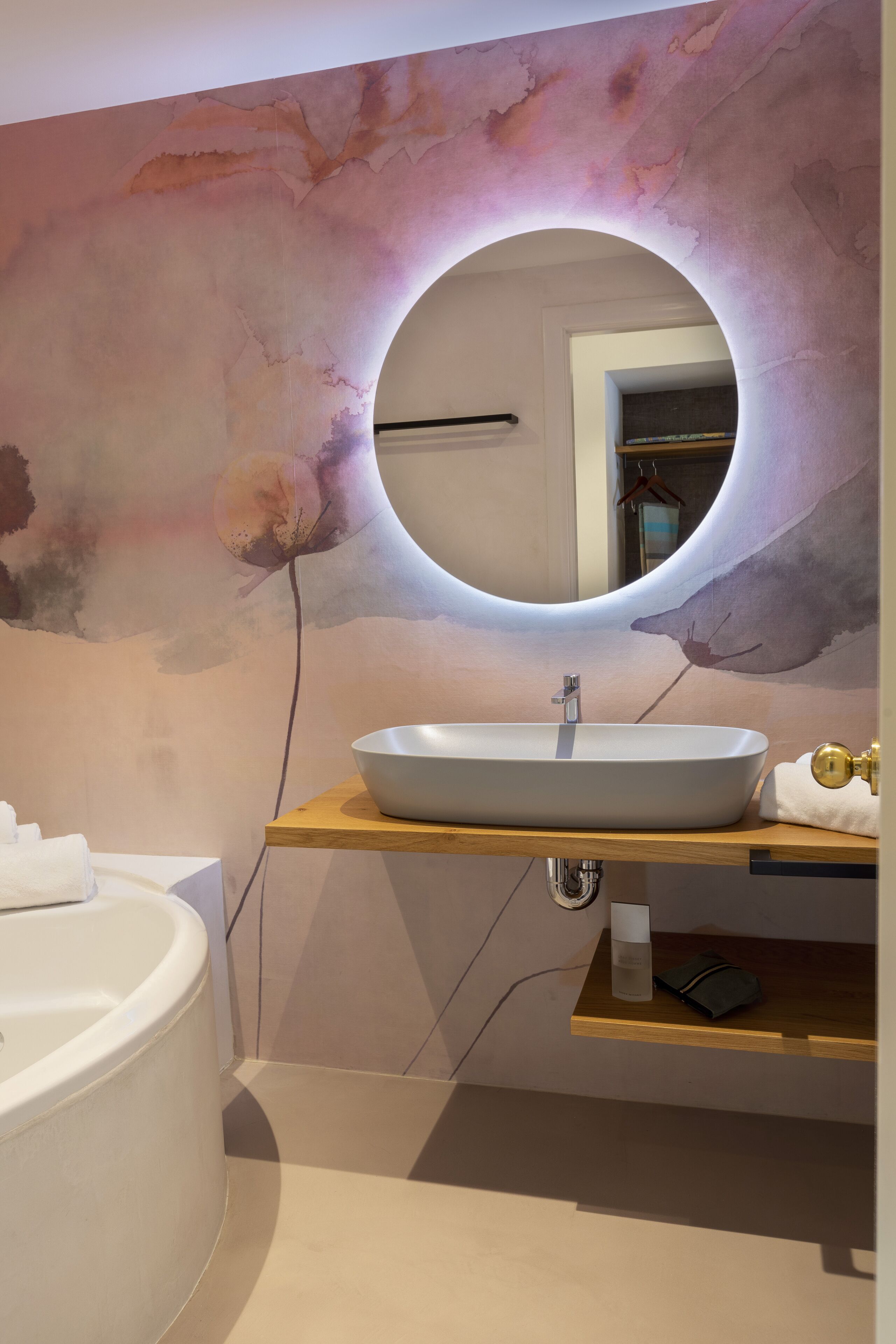 A modern bathroom with a white bathtub, a round mirror with a light halo, and a wooden vanity with a sink.