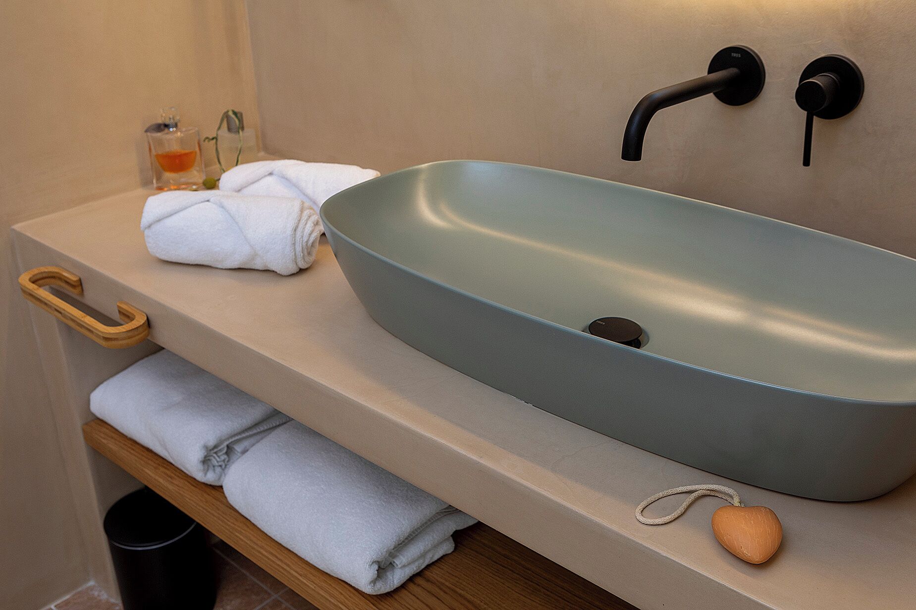 Un baño con un lavabo azul claro tipo vasija, dos anillos para toallas negros, un toallero de madera, un pequeño recipiente de vidrio con líquido amarillo y una pastilla de jabón con forma de corazón sobre el mostrador.
