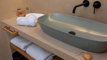 Un baño con un lavabo azul claro tipo vasija, dos anillos para toallas negros, un toallero de madera, un pequeño recipiente de vidrio con líquido amarillo y una pastilla de jabón con forma de corazón sobre el mostrador.