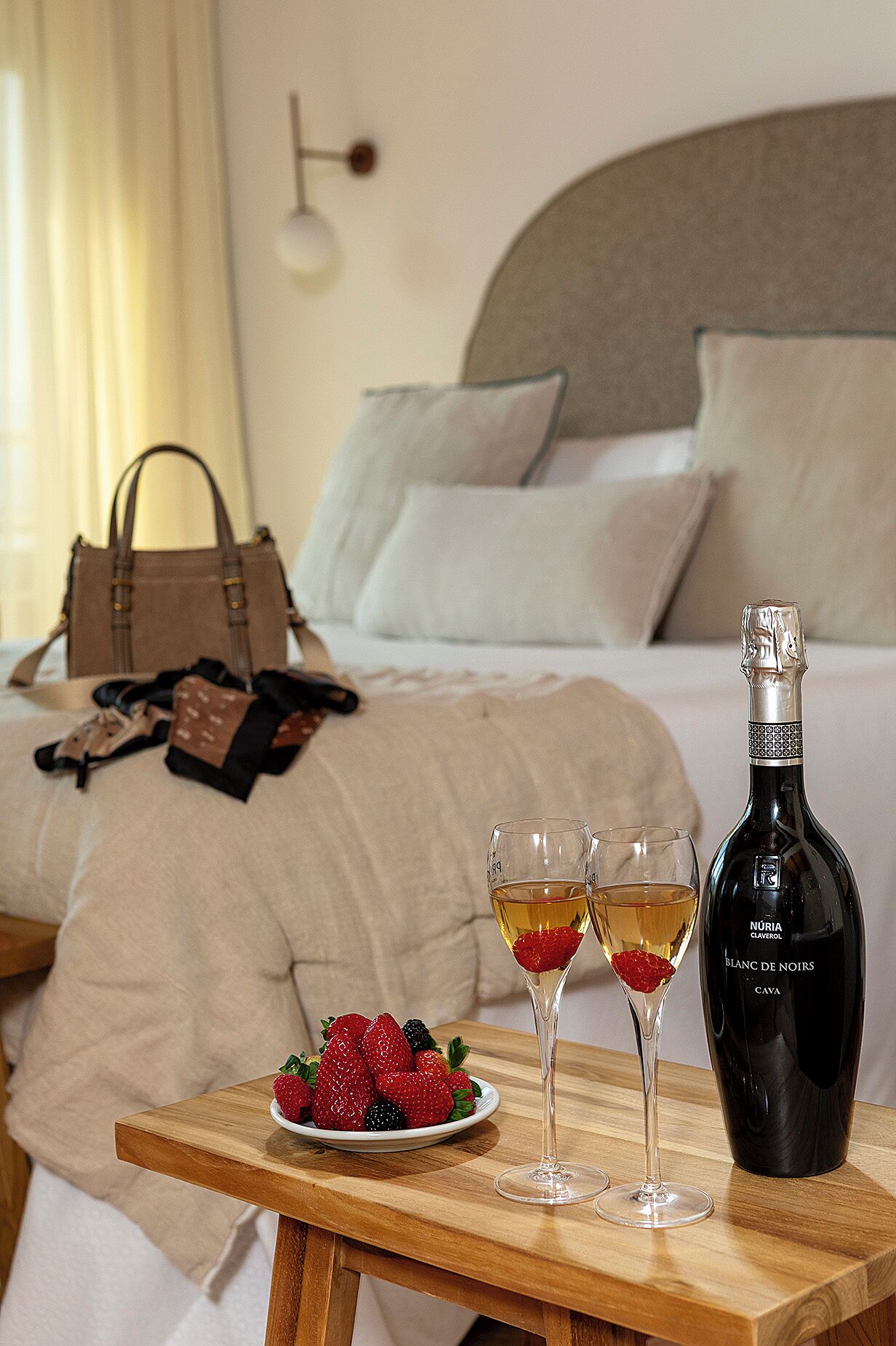 Ein ordentlich bezogenes Bett mit Kopfteil, ein Nachttisch mit einer Weinflasche, zwei Gläsern und einer Schale Erdbeeren und Brombeeren.