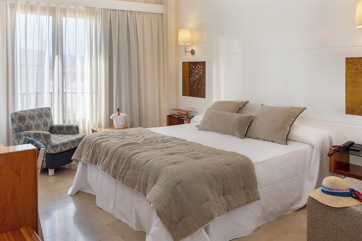 Une chambre d’hôtel avec un grand lit, une chaise, une table de chevet, une lampe et une vue sur l’extérieur à travers des rideaux légers.