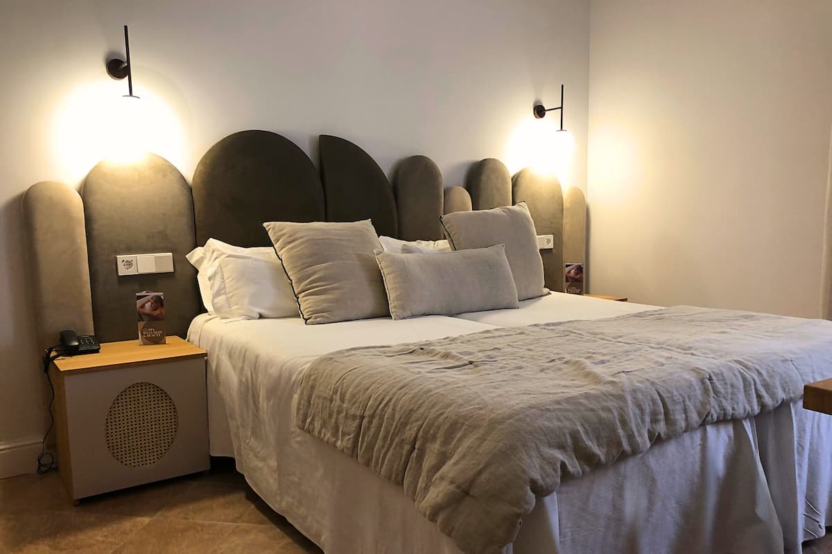Un lit bien fait, avec une couette blanche et plusieurs oreillers, une table de chevet sur laquelle se trouvent un téléphone et un petit objet décoratif, ainsi que des luminaires fixés au mur.