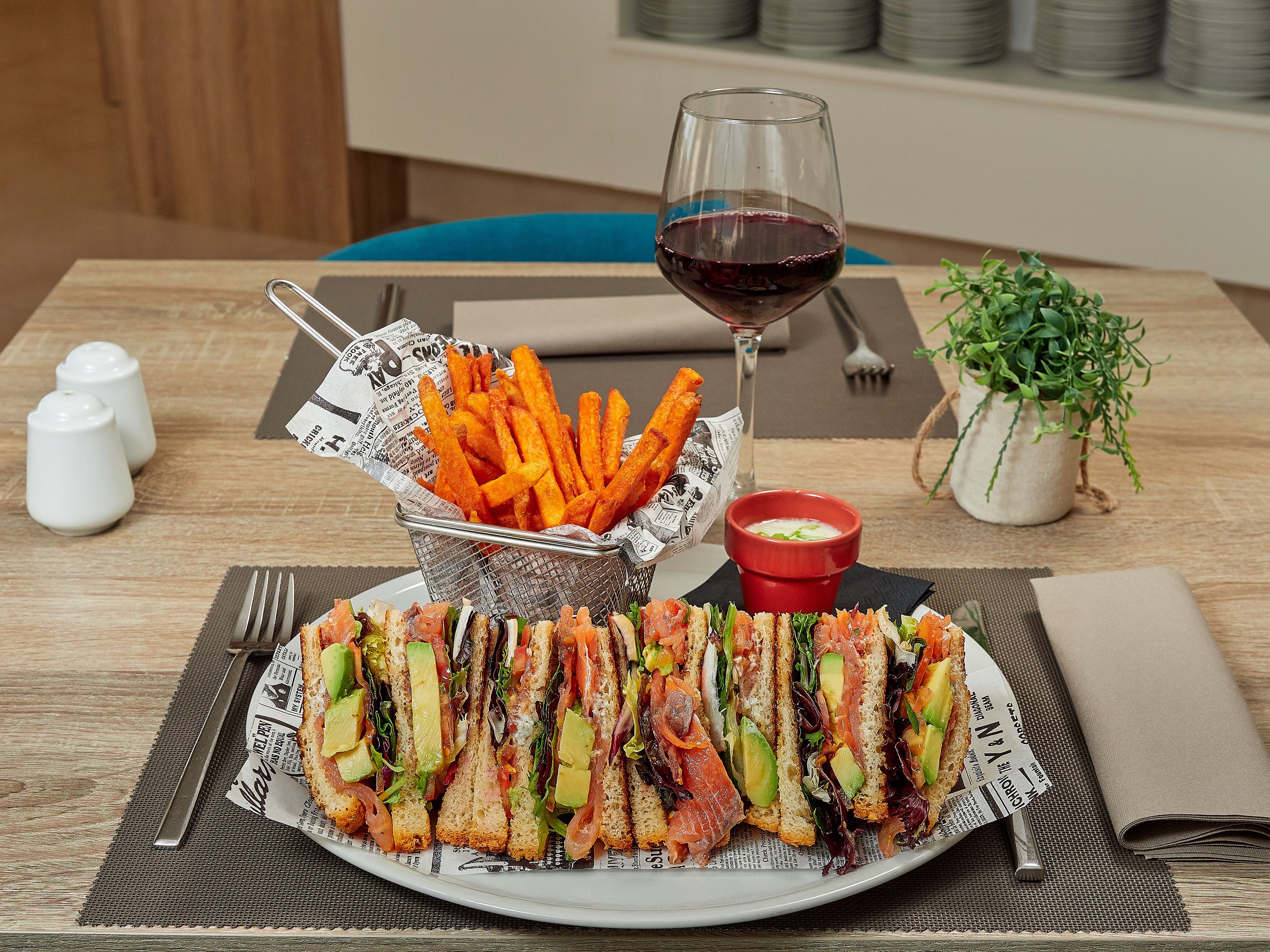 A plate of sandwiches, a basket of fries, a glass of red wine, and a small bowl of sauce on a wooden table.