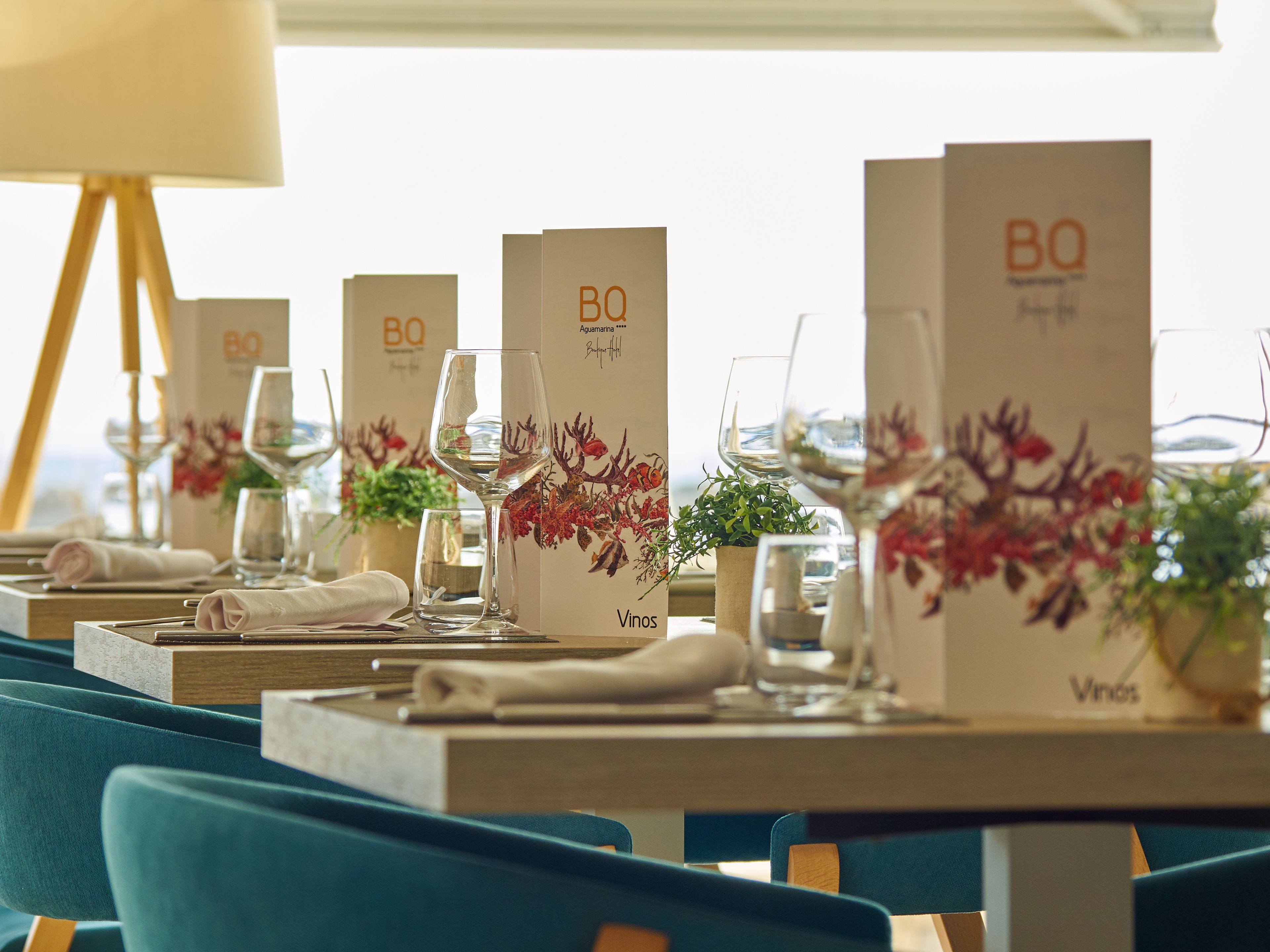 A restaurant setting with tables set for dining, each with a menu stand, wine glasses, and decorative plants.