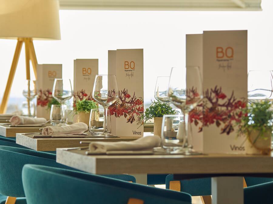 A restaurant setting with tables set for dining, each with a menu stand, wine glasses, and decorative plants.