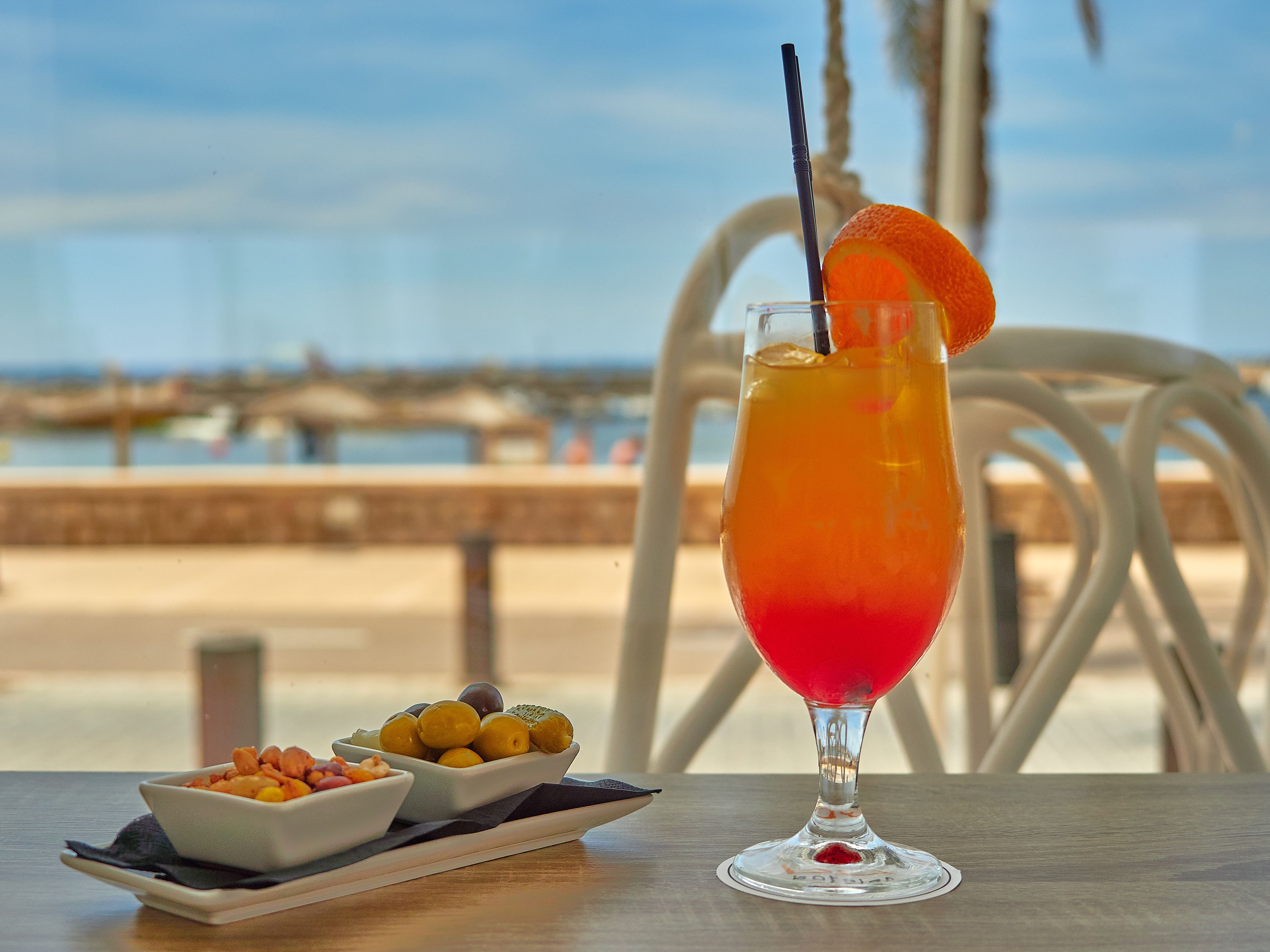 Una mesa con un vaso de bebida roja, un cuenco de fruta y vista al mar.