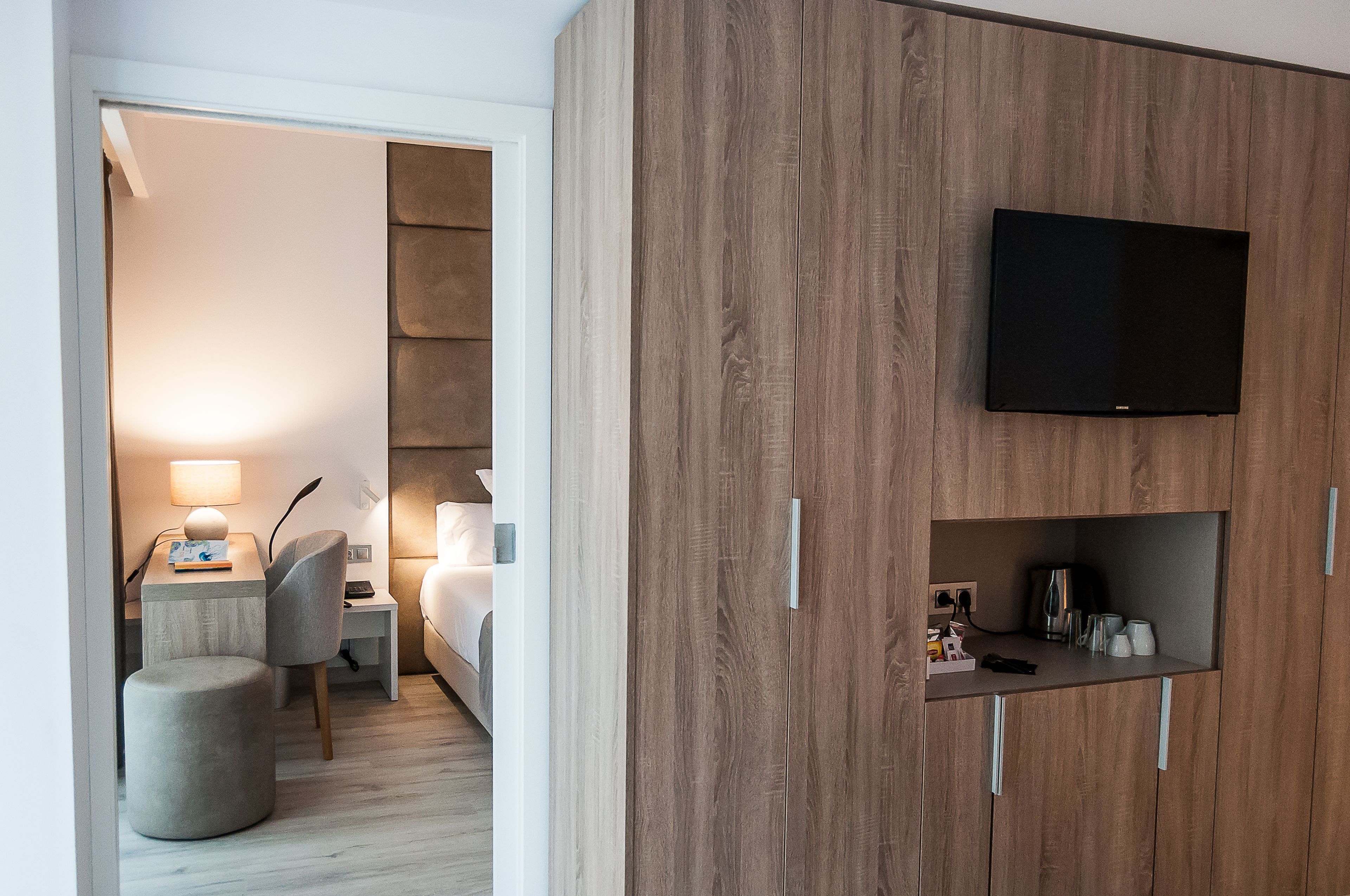 Una habitación de hotel moderna con un armario de madera, un televisor de pantalla plana montado encima, una mesita de noche con una lámpara, una silla y un pequeño reposapiés.