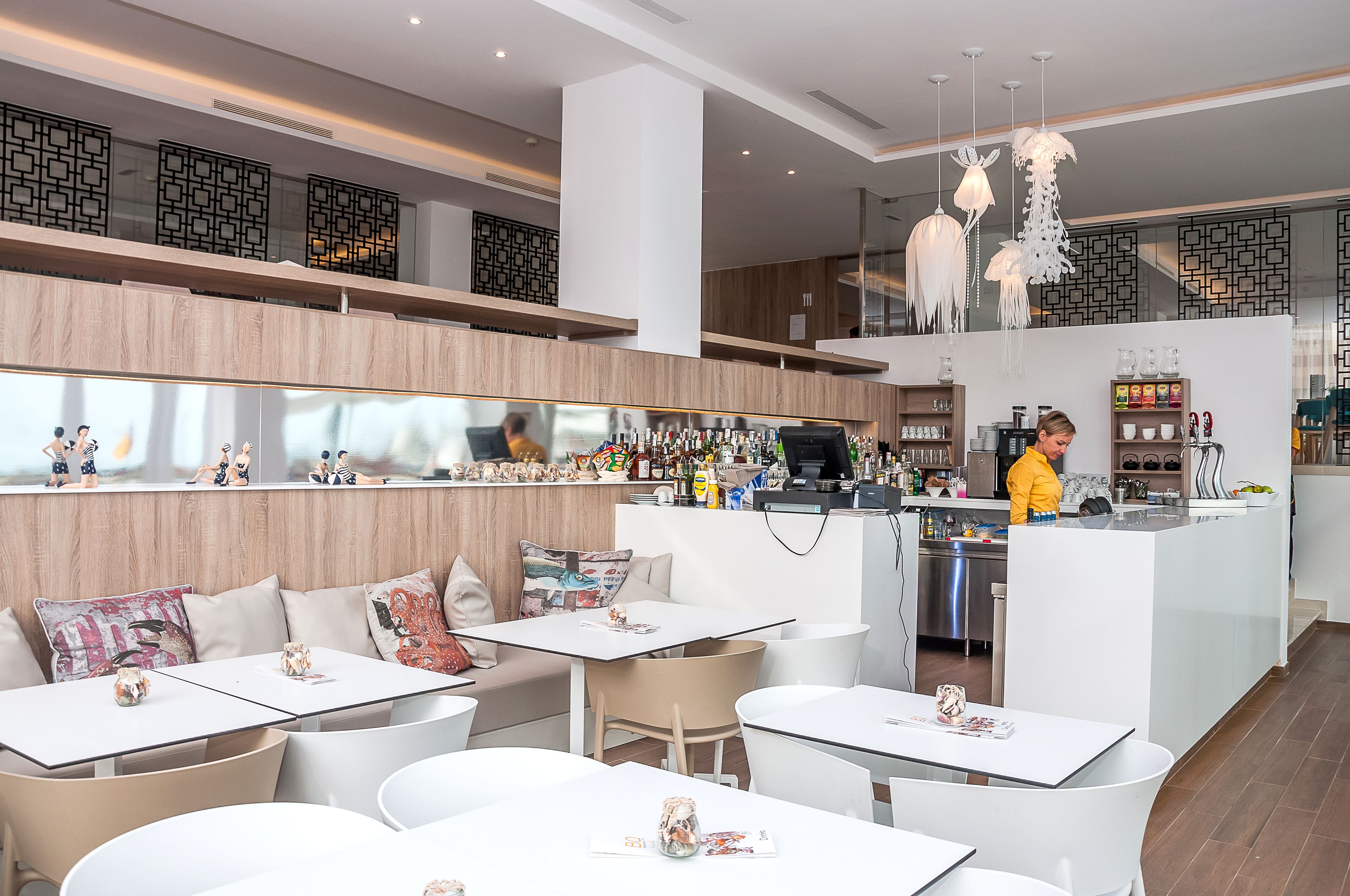 Un restaurante moderno con mobiliario blanco, una zona de bar y una cocina visible al fondo.