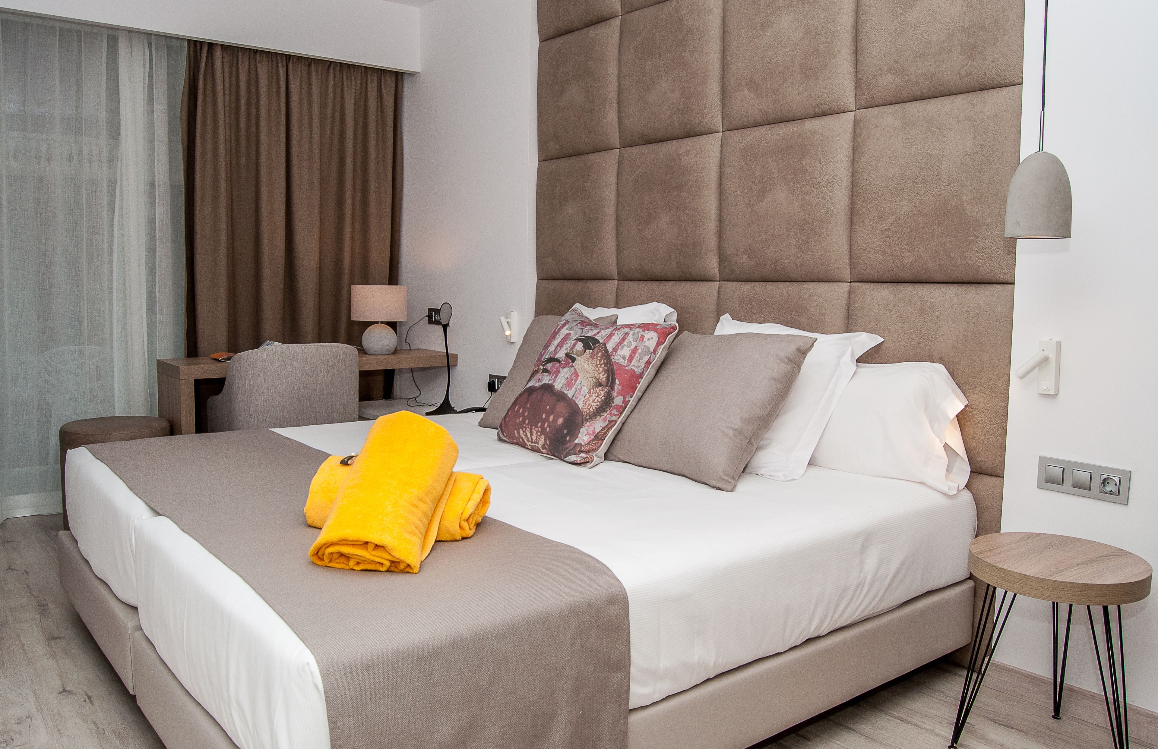 A modern hotel room with a large bed, a bedside table with a lamp, a small round table, and a large window with curtains.