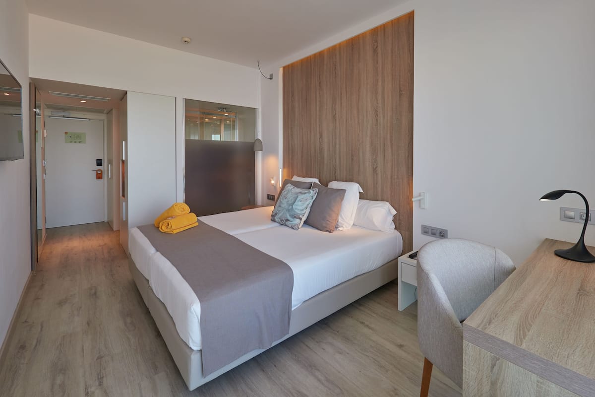 A modern hotel room with a neatly made bed, a desk with a lamp, and a wooden accent wall.