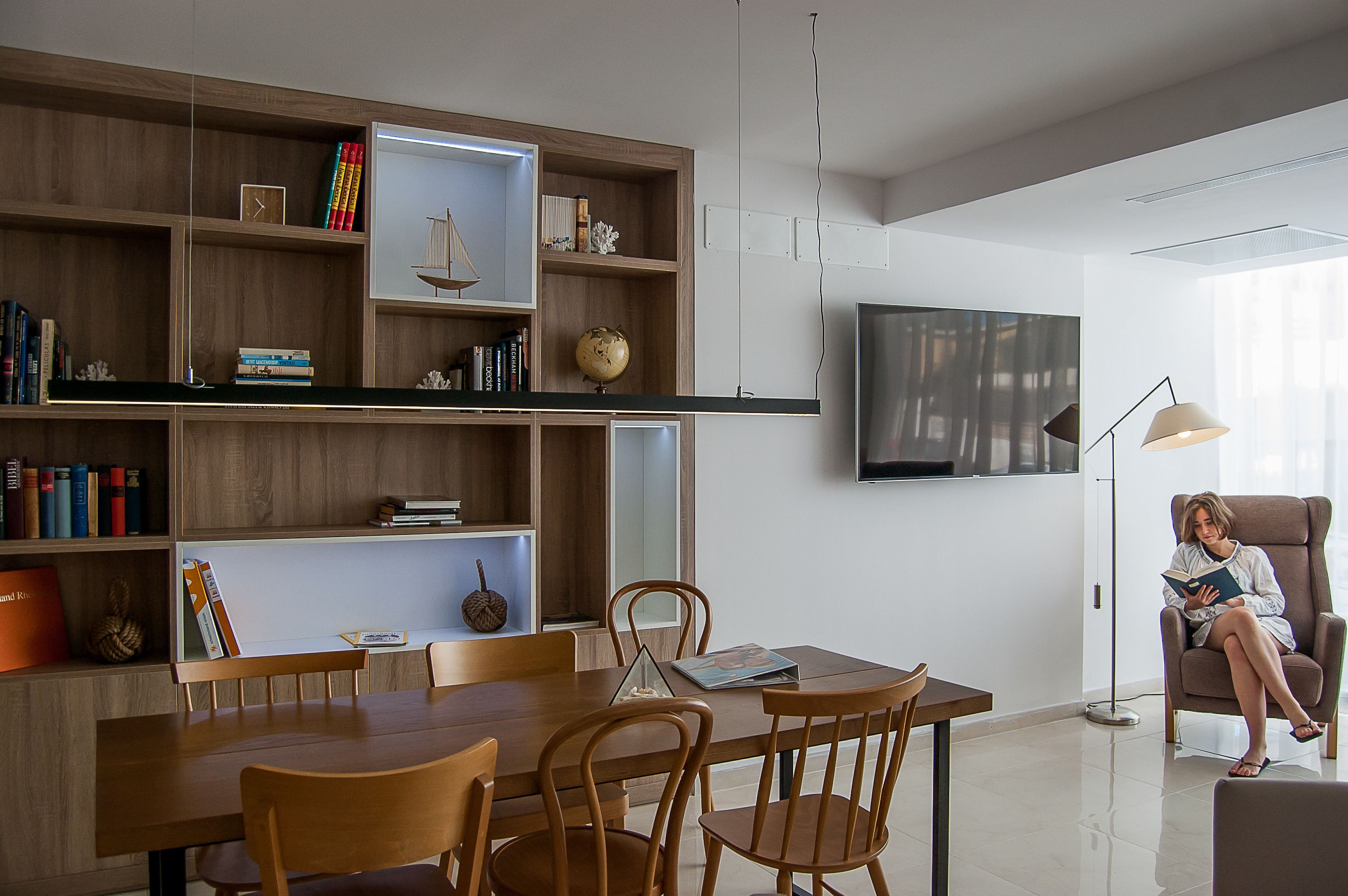 Una sala de estar moderna con comedor, muebles de madera, un televisor de pantalla plana y una persona leyendo un libro.