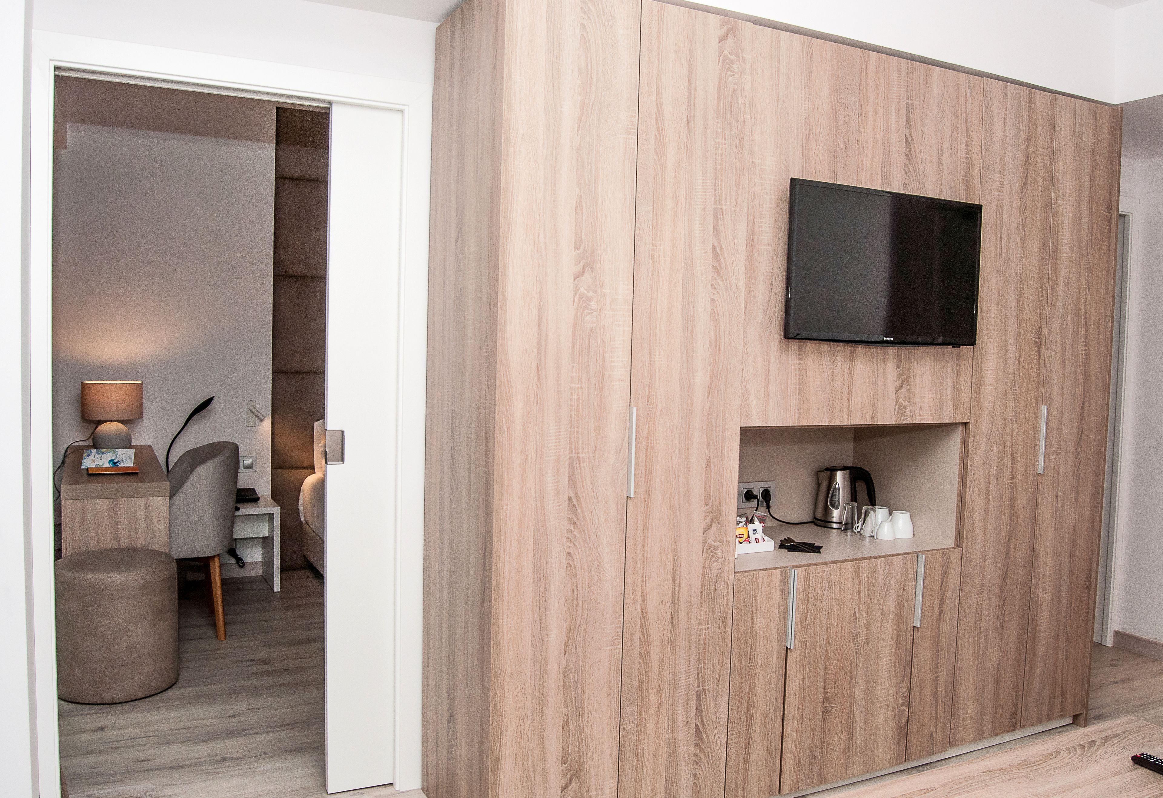 Quarto moderno com TV integrada, pequena cozinha e área de estar com luminária e poltrona.