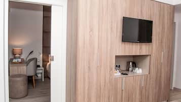 Quarto moderno com TV integrada, pequena cozinha e área de estar com luminária e poltrona.