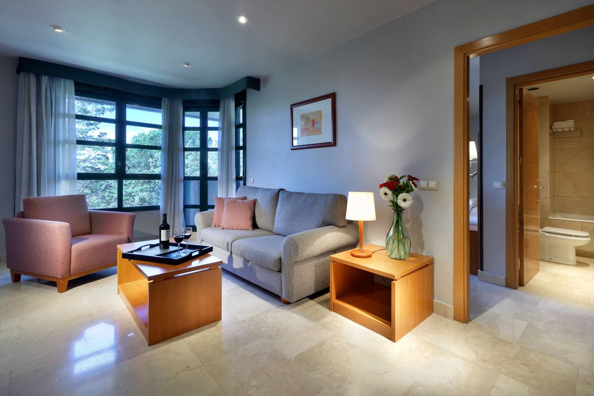 Una sala de estar con sofá, sillón, mesa de centro y una mesita auxiliar con lámpara y flores.