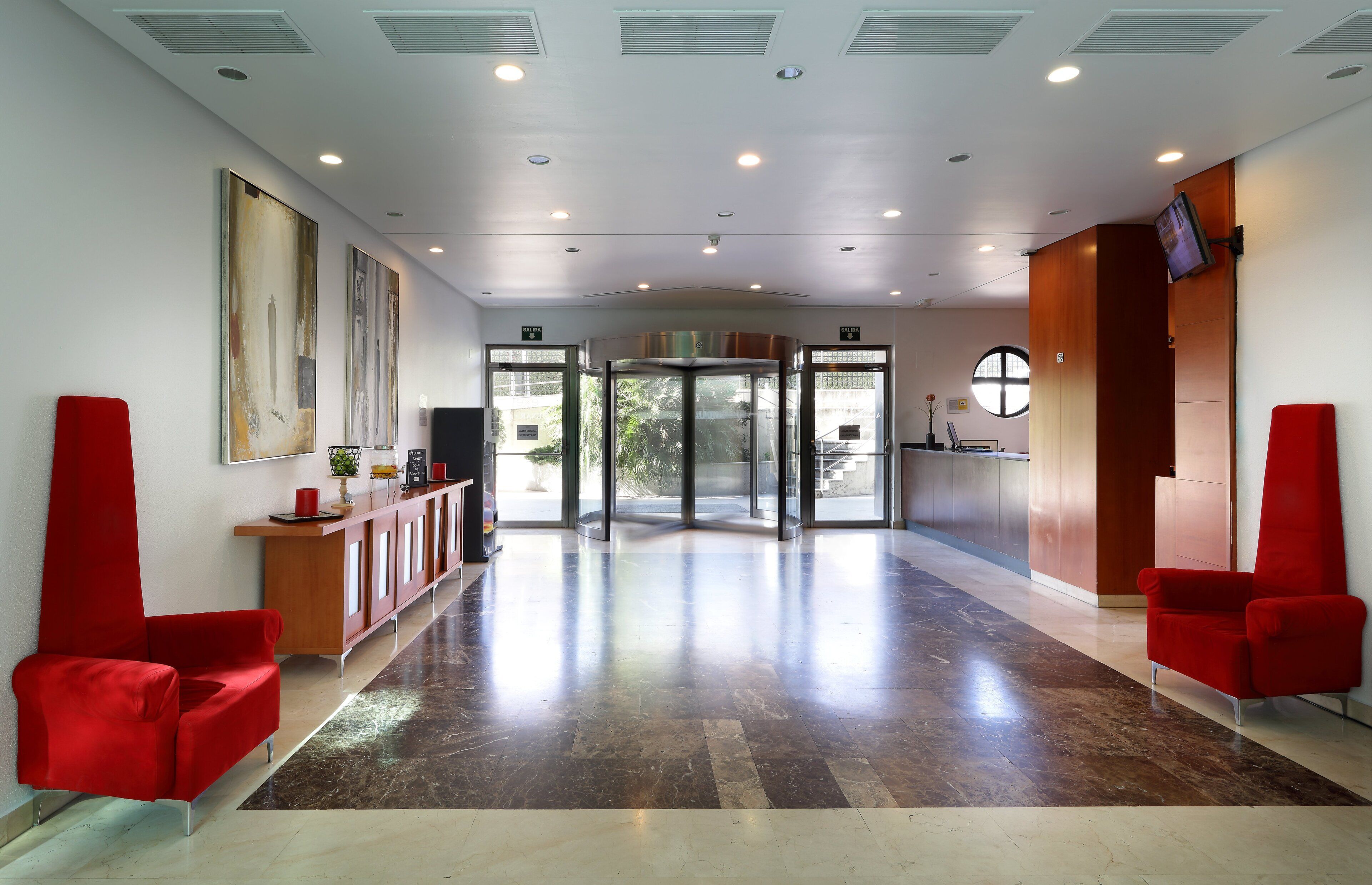 A lobby with a reception desk, red chairs, a revolving door, and a large window.