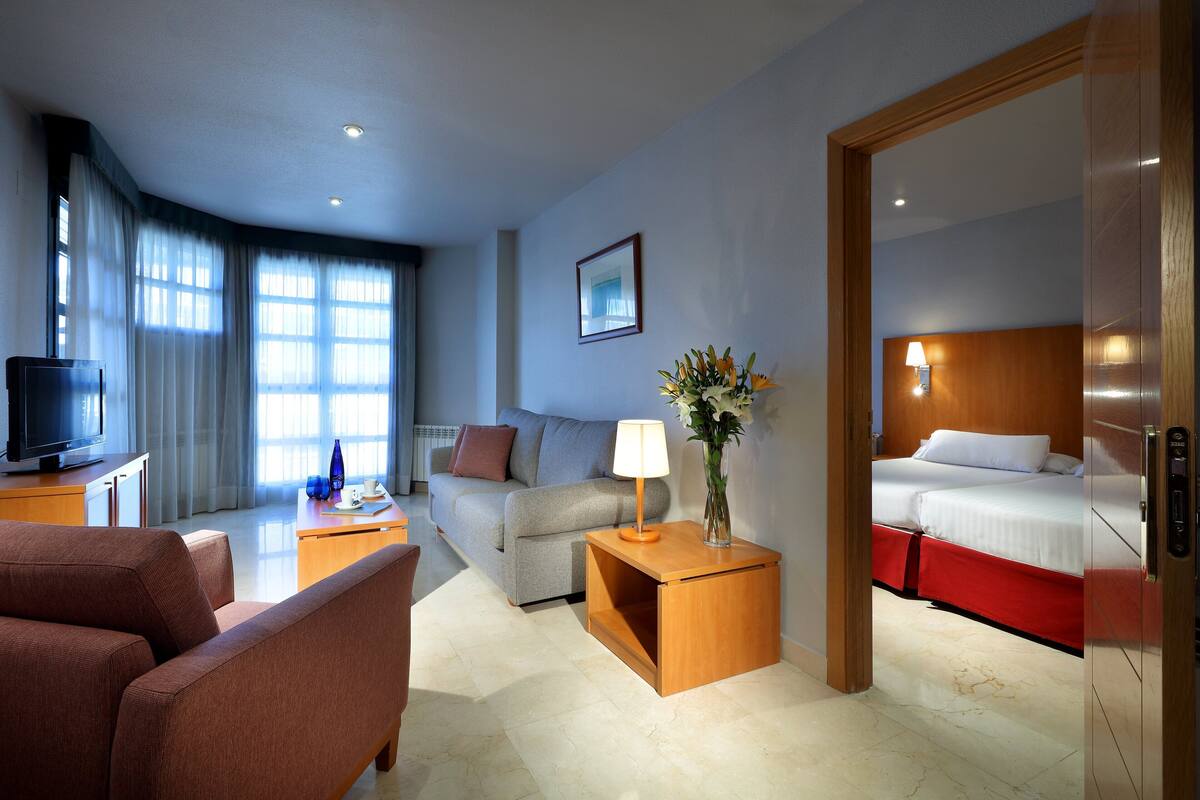 Habitación de hotel con sofá, una pequeña mesa con lámpara, televisor, cama con ropa de cama roja y un espejo.
