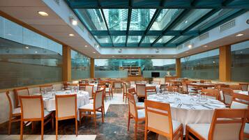 Un comedor amplio con varias mesas dispuestas para una comida, ventanas grandes y un techo alto con paneles de vidrio.