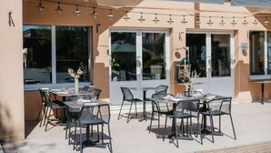 An outdoor cafe with tables and chairs, string lights, and large windows.