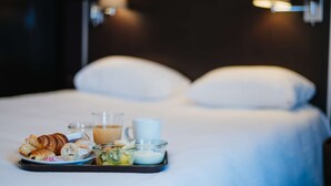 A neatly made bed with white linens and two wall-mounted lamps. A breakfast tray with croissants, a glass of orange juice, and a cup is placed on the bed.