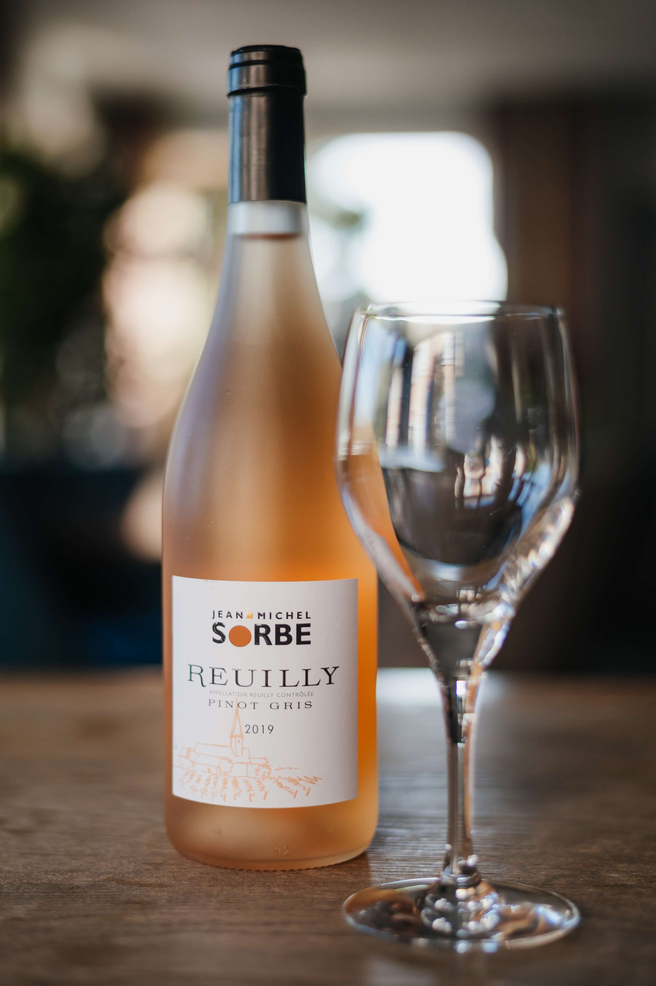 Egy üveg Jean Michel Sorbe Reuilly Pinot Gris 2019-es évjáratú bor egy üres borospohár mellett, egy fafelületen.