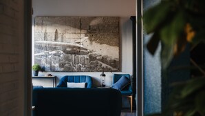 A room with a large cityscape painting on the wall, a blue sofa, a small table with a lamp, and a potted plant.
