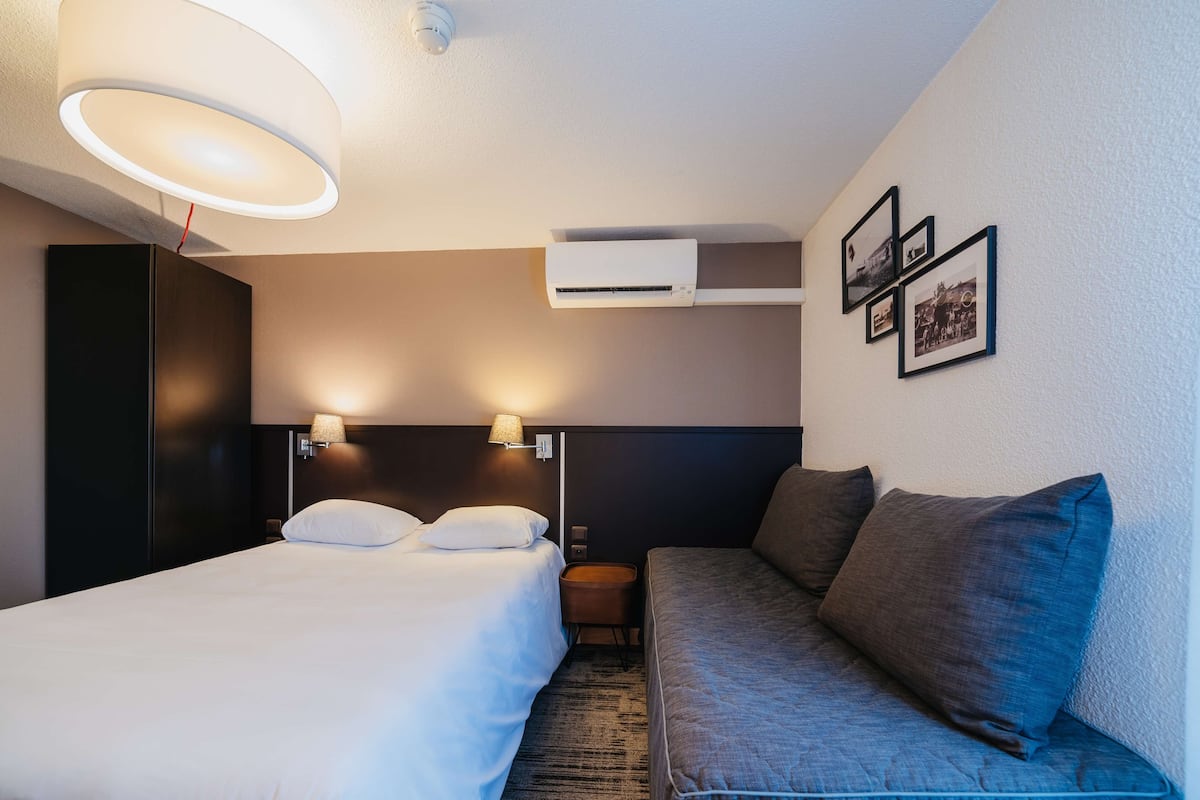 A hotel room with a bed, a sofa, a wall-mounted air conditioner, and framed pictures on the wall.
