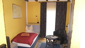 Quarto pequeno com cama, escrivaninha, cadeira e janela com cortinas.