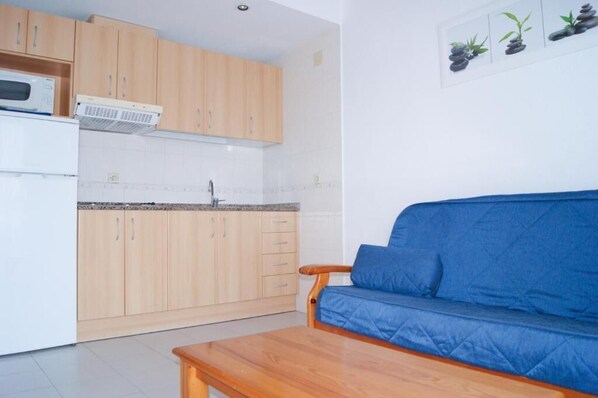 A compact kitchen with wooden cabinets, a microwave, and a sink. There is a blue sofa and a wooden table in the living area. - Apartamentos Palmera Beach (Orihuela)