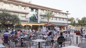 Un área de comedor al aire libre con varias mesas y sillas, un edificio con balcones y una palmera.