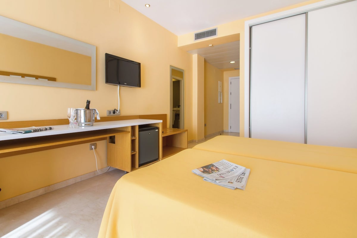 Habitación de hotel con cama, escritorio con televisor, pequeña kitchenette y puerta que da a otra habitación.