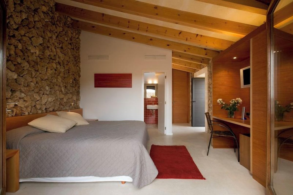 A bedroom with a stone wall, a bed with a grey bedspread, a red rug, and a wooden desk with a chair.