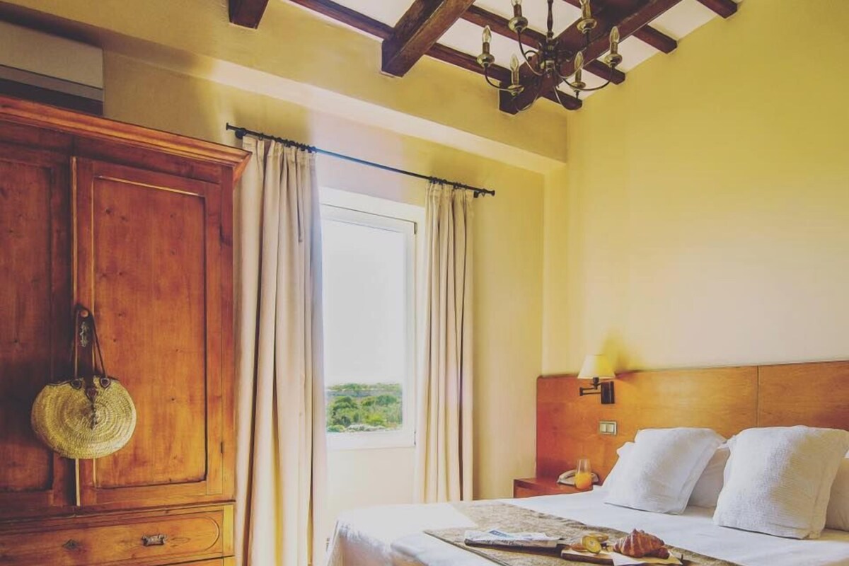 Un dormitorio con un armario de madera, una cama con ropa de cama blanca, una mesita de noche con lámpara y vistas a la vegetación por la ventana.
