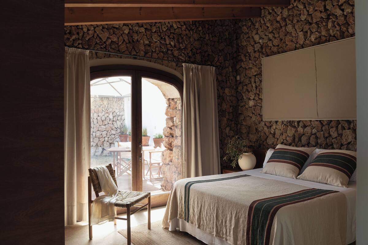 Une chambre avec un mur en pierre, un lit agrémenté d’oreillers à rayures, une chaise et une vue sur l’extérieur, accessible par une porte vitrée.