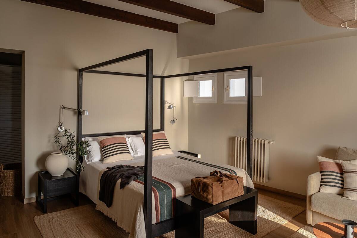 Un lit à baldaquin avec une structure sombre, une table de chevet avec une lampe, un fauteuil avec des coussins et un radiateur fixé au mur.