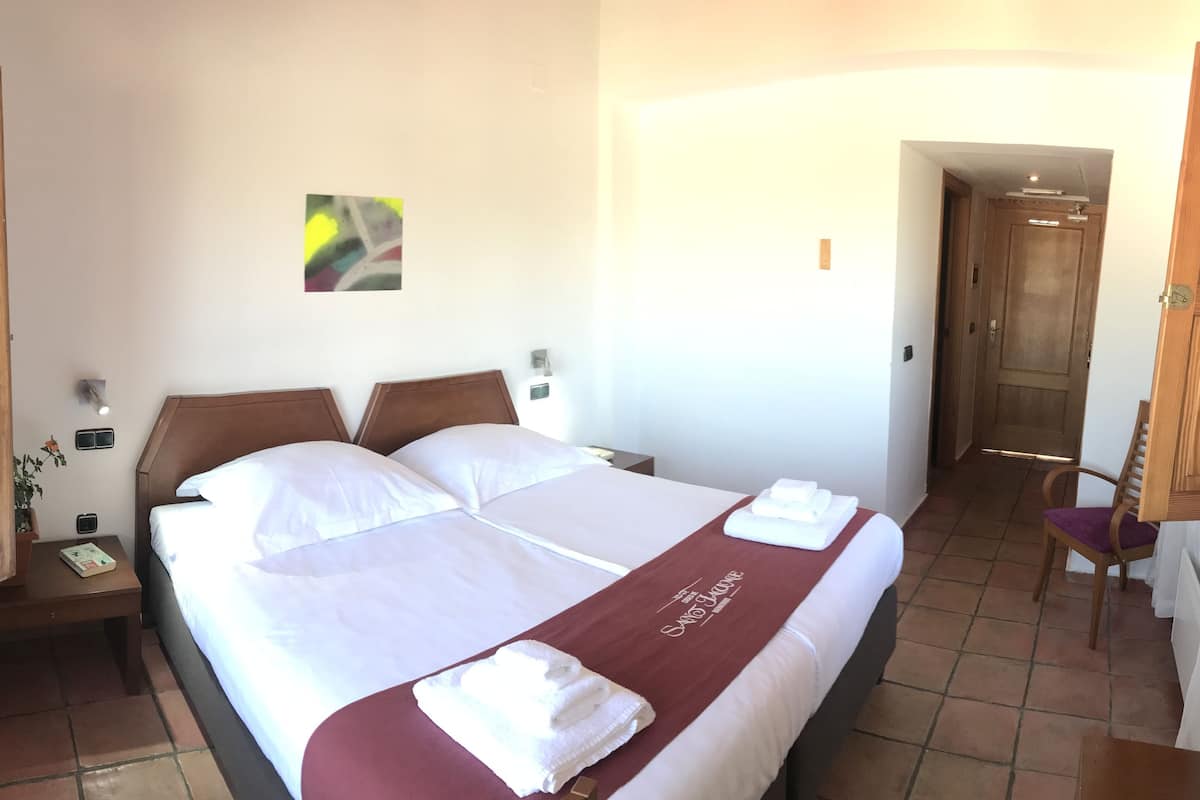 Une chambre d’hôtel avec un lit double, une tête de lit en bois, une table de chevet avec une lampe, une petite plante en pot et une œuvre d’art accrochée au mur.