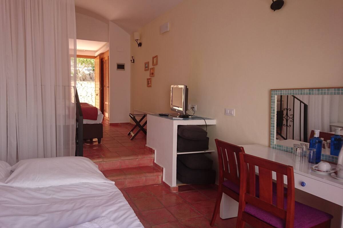 Une chambre à coucher avec un lit, un bureau et une chaise. Il y a une fenêtre avec des rideaux, un miroir et une petite table sur laquelle sont posées des bouteilles.