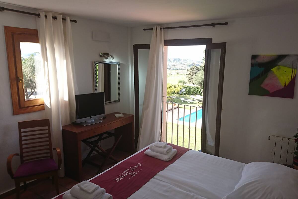Une chambre d’hôtel avec un lit, un bureau avec une télévision, une chaise, un balcon donnant sur une piscine et un tableau au mur.