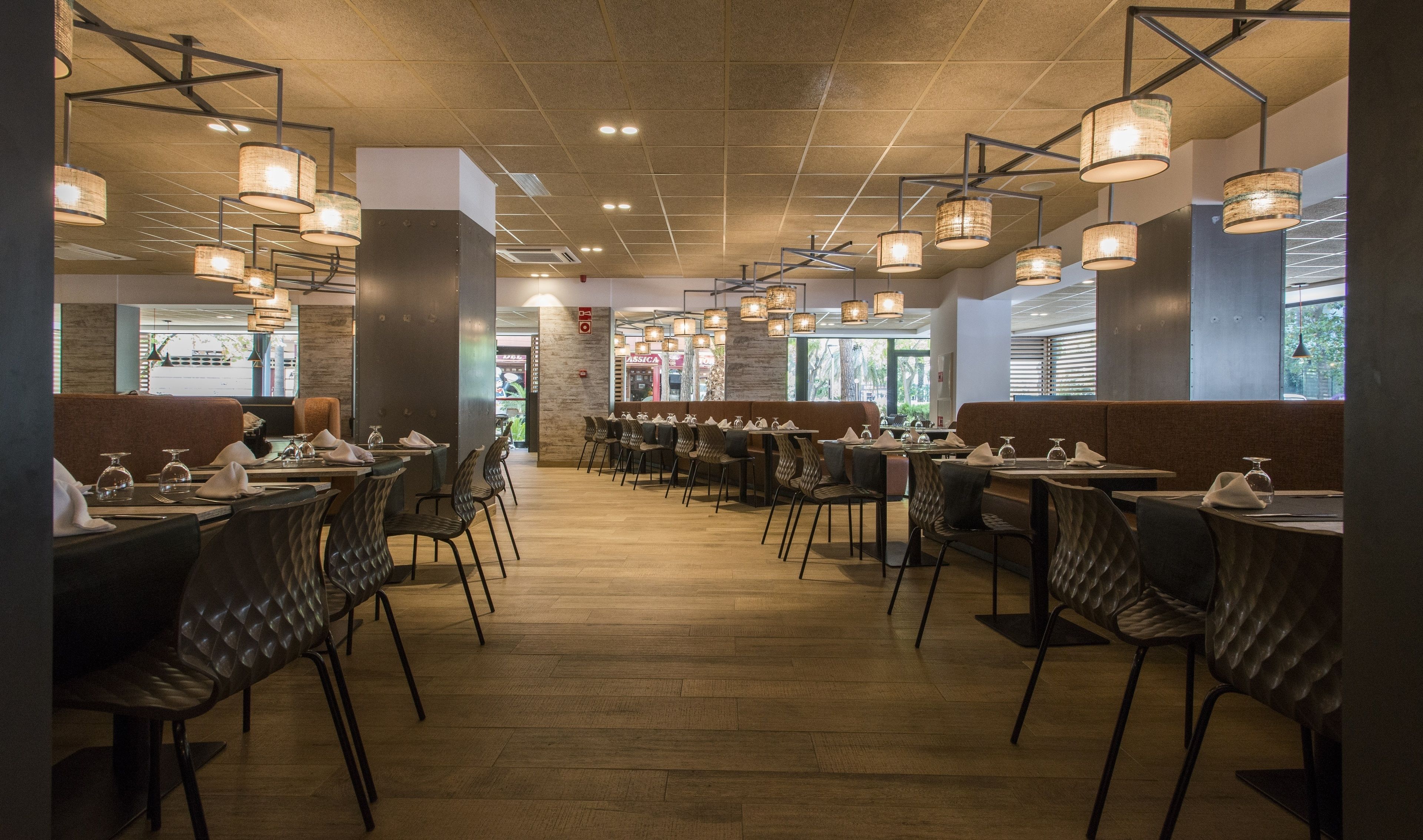 A restaurant with a modern interior, featuring wooden flooring, black upholstered chairs, and hanging pendant lights. Tables are set with white tablecloths and napkins.