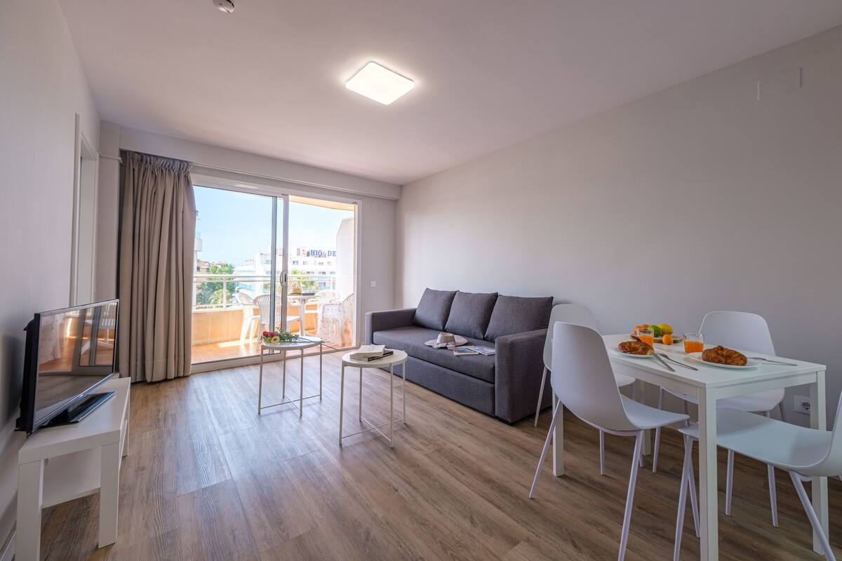A modern living room with a grey sofa, a flat-screen TV, a dining table with chairs, and a balcony with a view of the city.