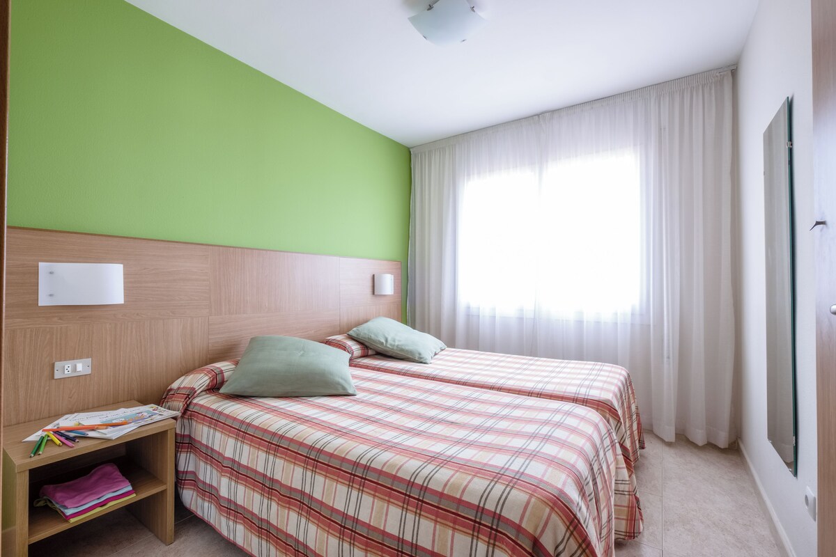 Une chambre avec deux lits, une tête de lit en bois, une table de chevet avec une lampe et une fenêtre avec des rideaux.