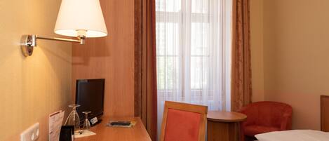 A hotel room with a wooden desk, a television, a bed, a chair, and a window with curtains.