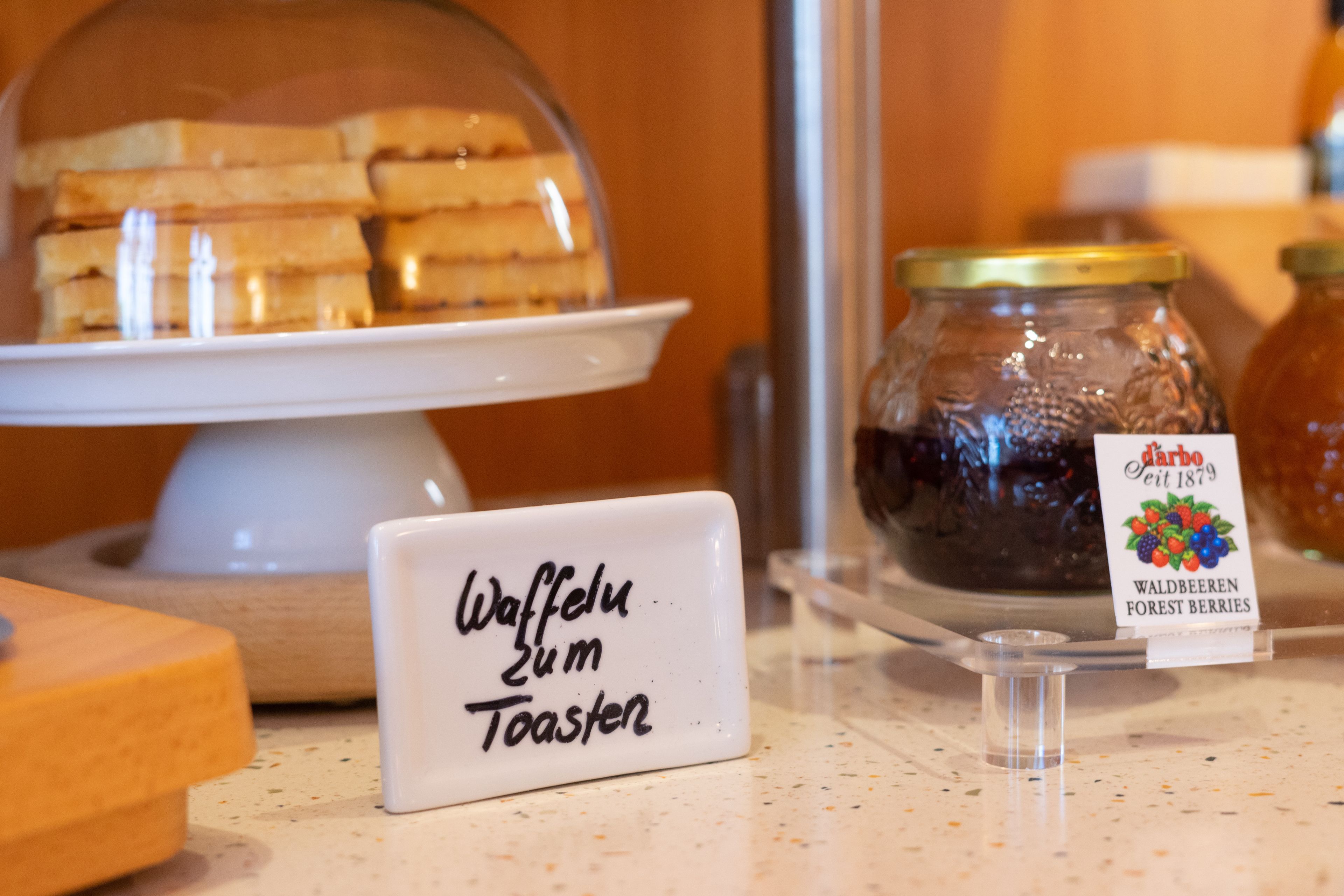 A sign indicating a waffle with toast option, a glass jar of forest berries, and a display of sandwiches under a dome.