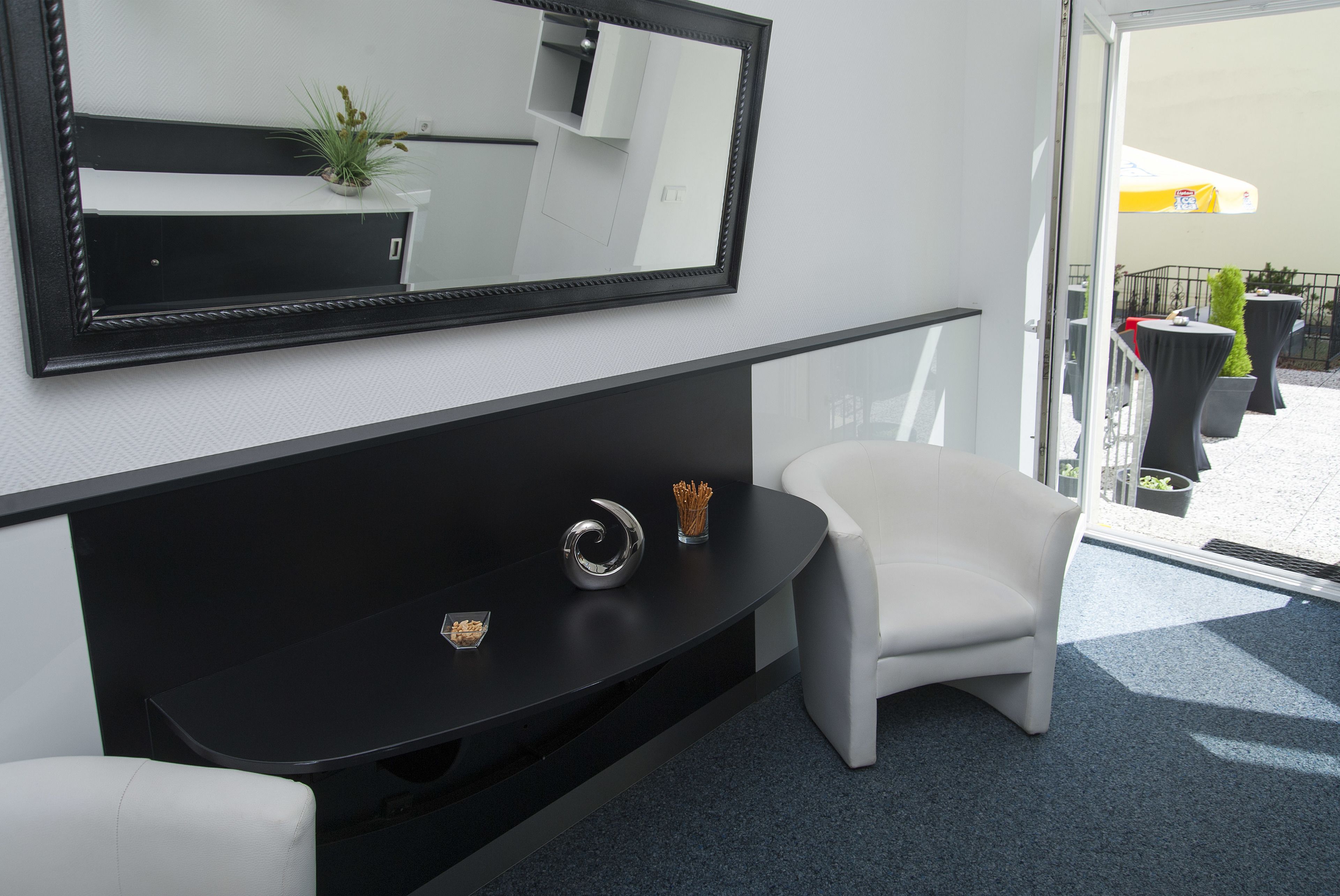 A modern hotel room with a black console table, a white armchair, a large mirror, and a balcony with outdoor seating.