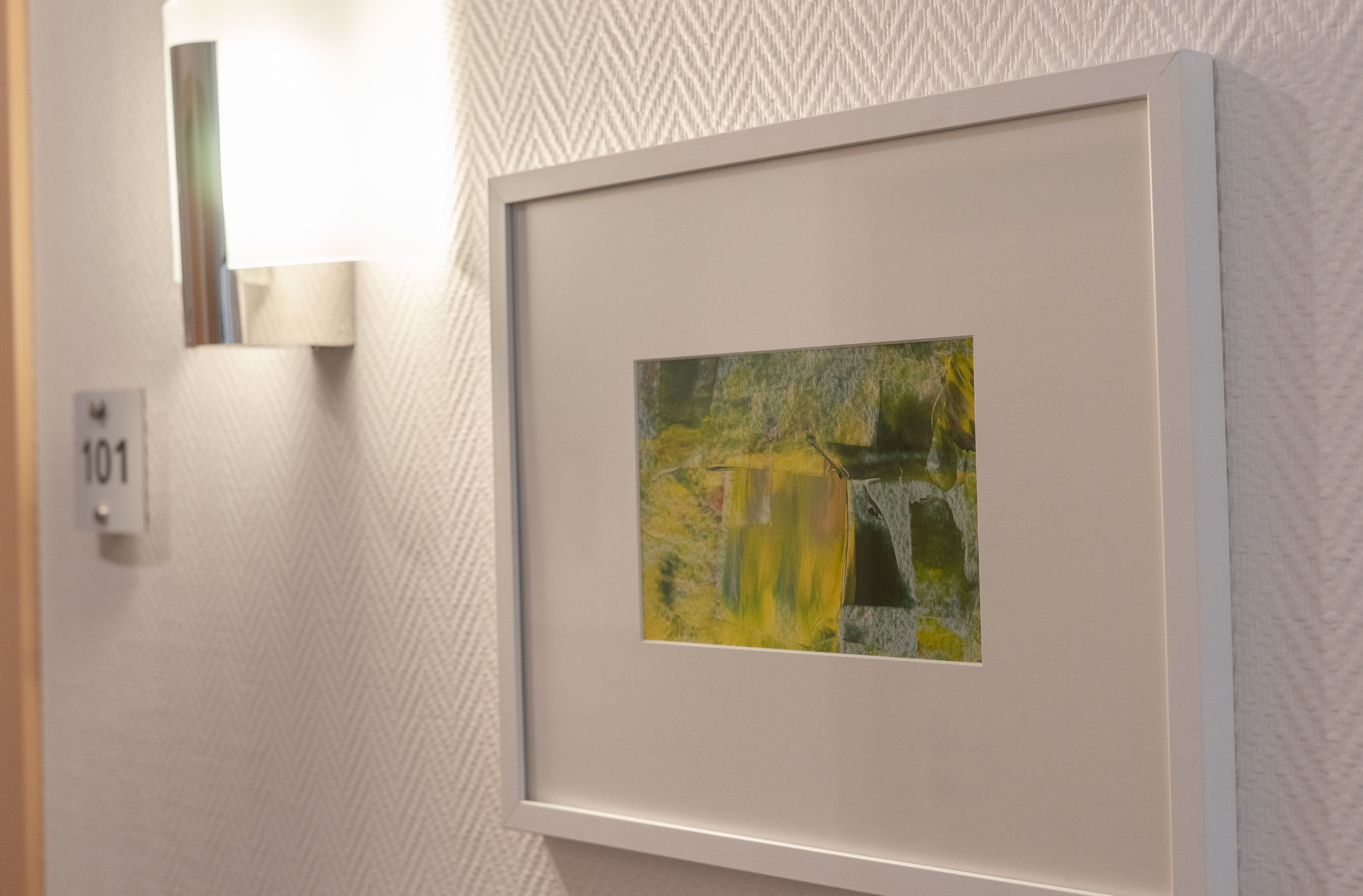 A framed artwork on a textured wall, a wall-mounted light fixture, and a door with a number plate.