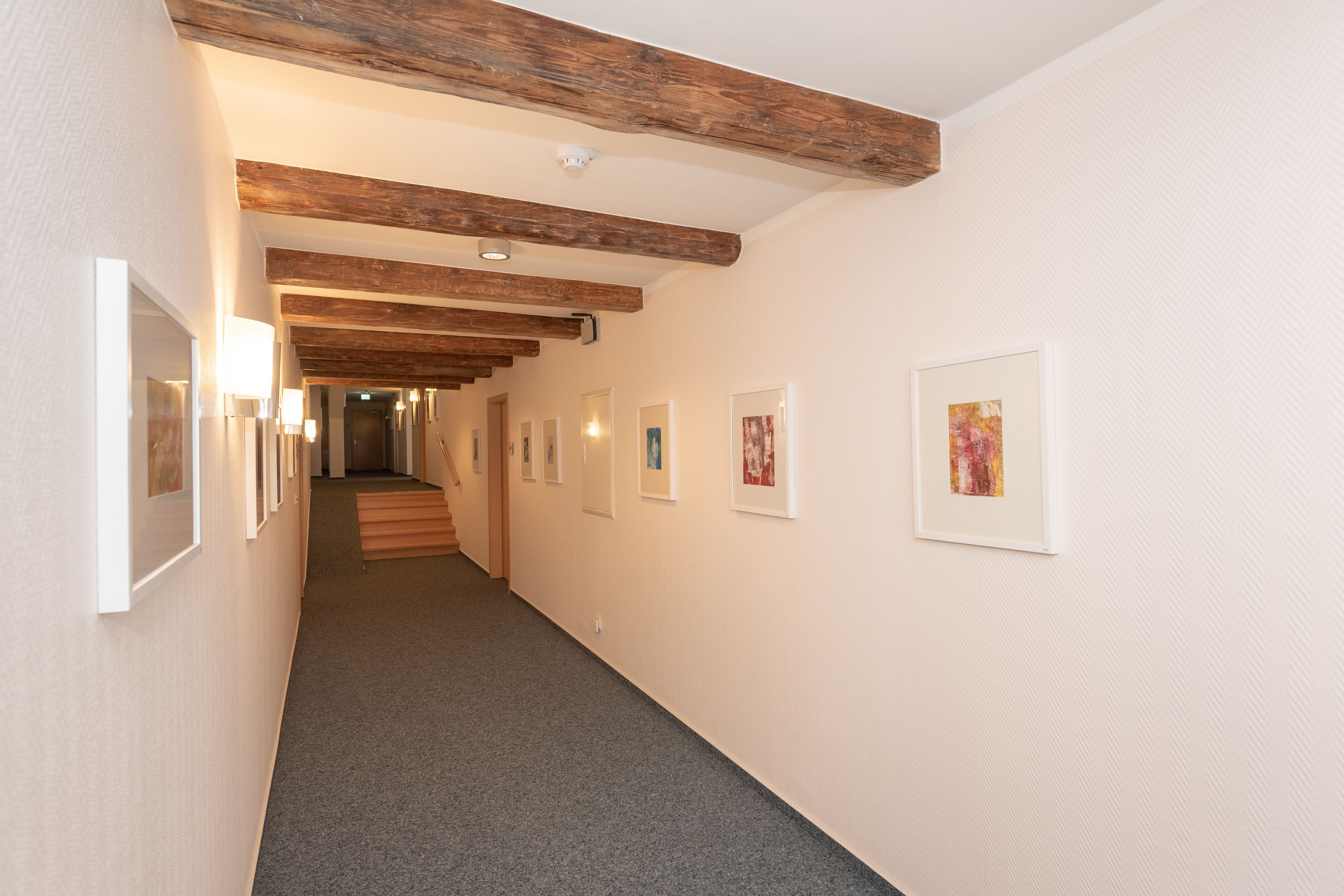 A hallway with framed artwork on the walls, wooden beams on the ceiling, and carpeted flooring.