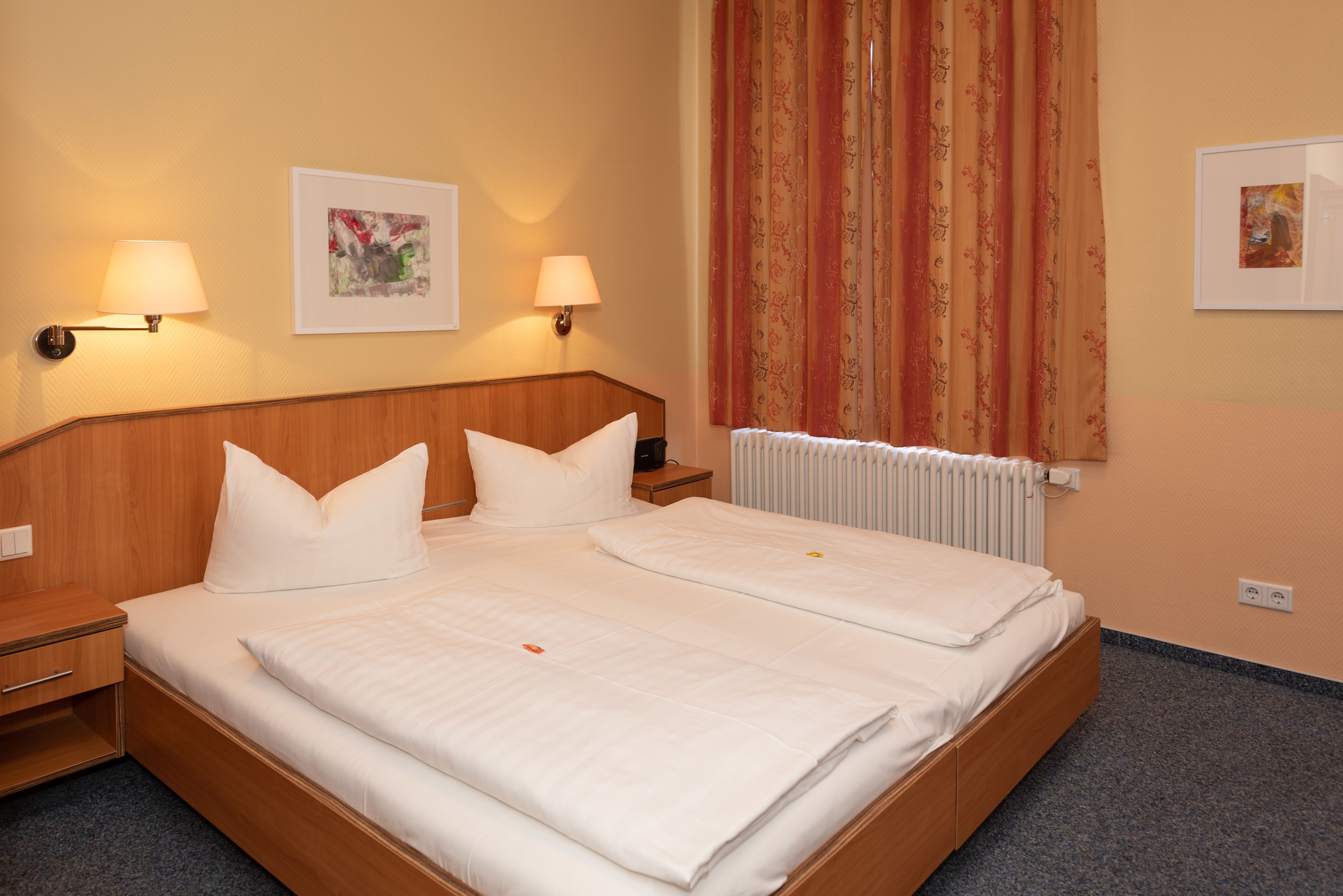 Une chambre d’hôtel avec un grand lit, deux tables de chevet, deux lampes fixées au mur et deux œuvres d’art encadrées.