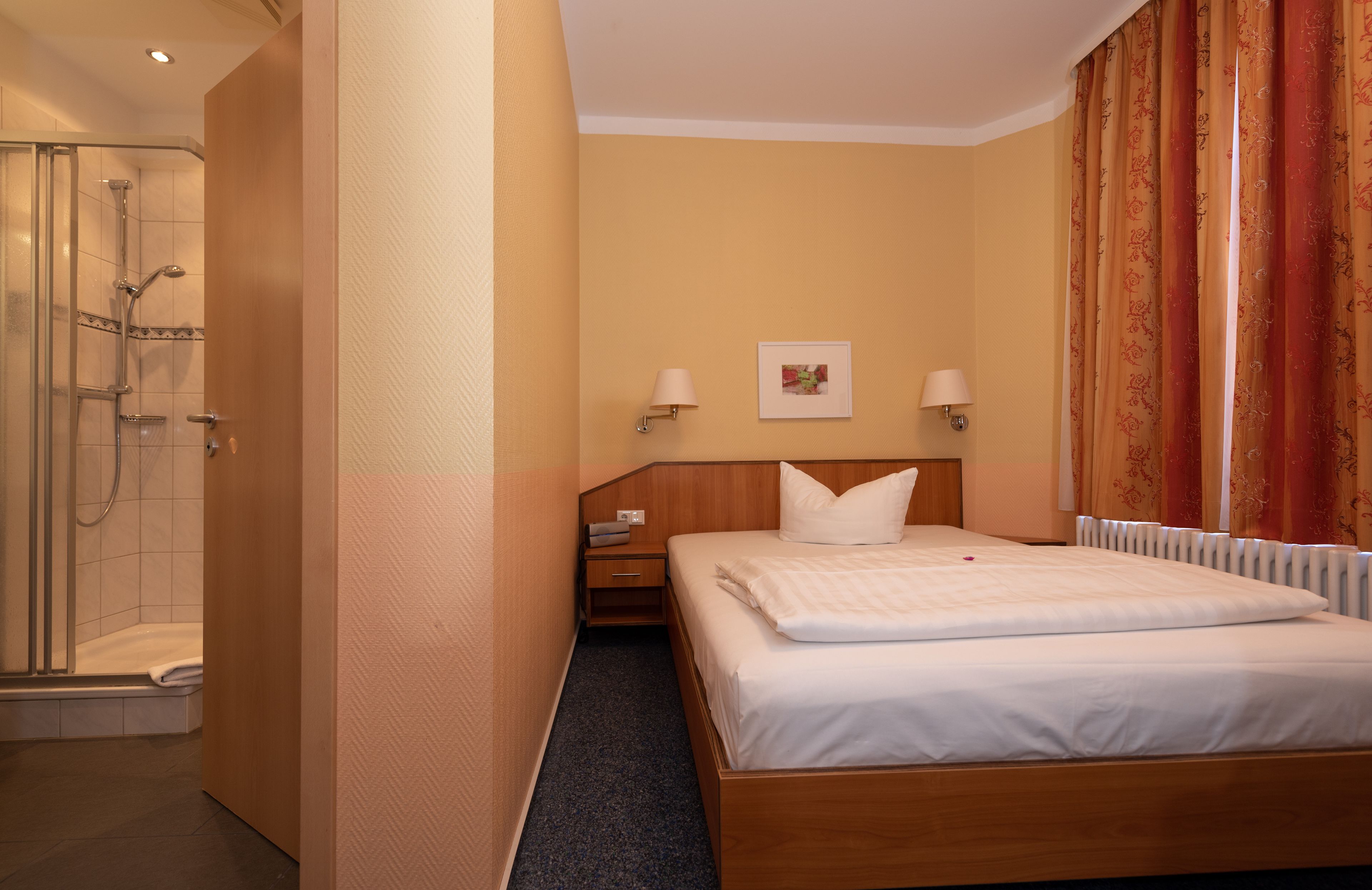 A hotel room with a shower, a bed, a nightstand, and a wall-mounted lamp.