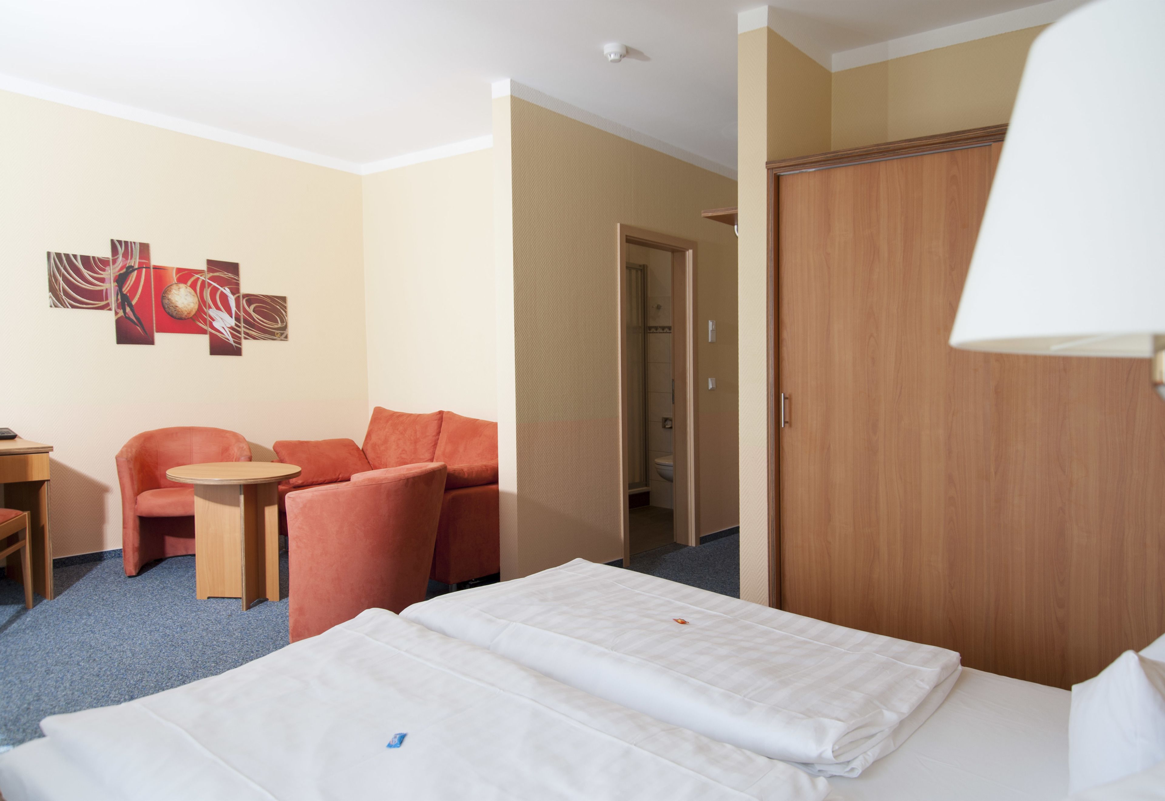A hotel room with a bed, a desk, a chair, a small table, and a door leading to a bathroom.