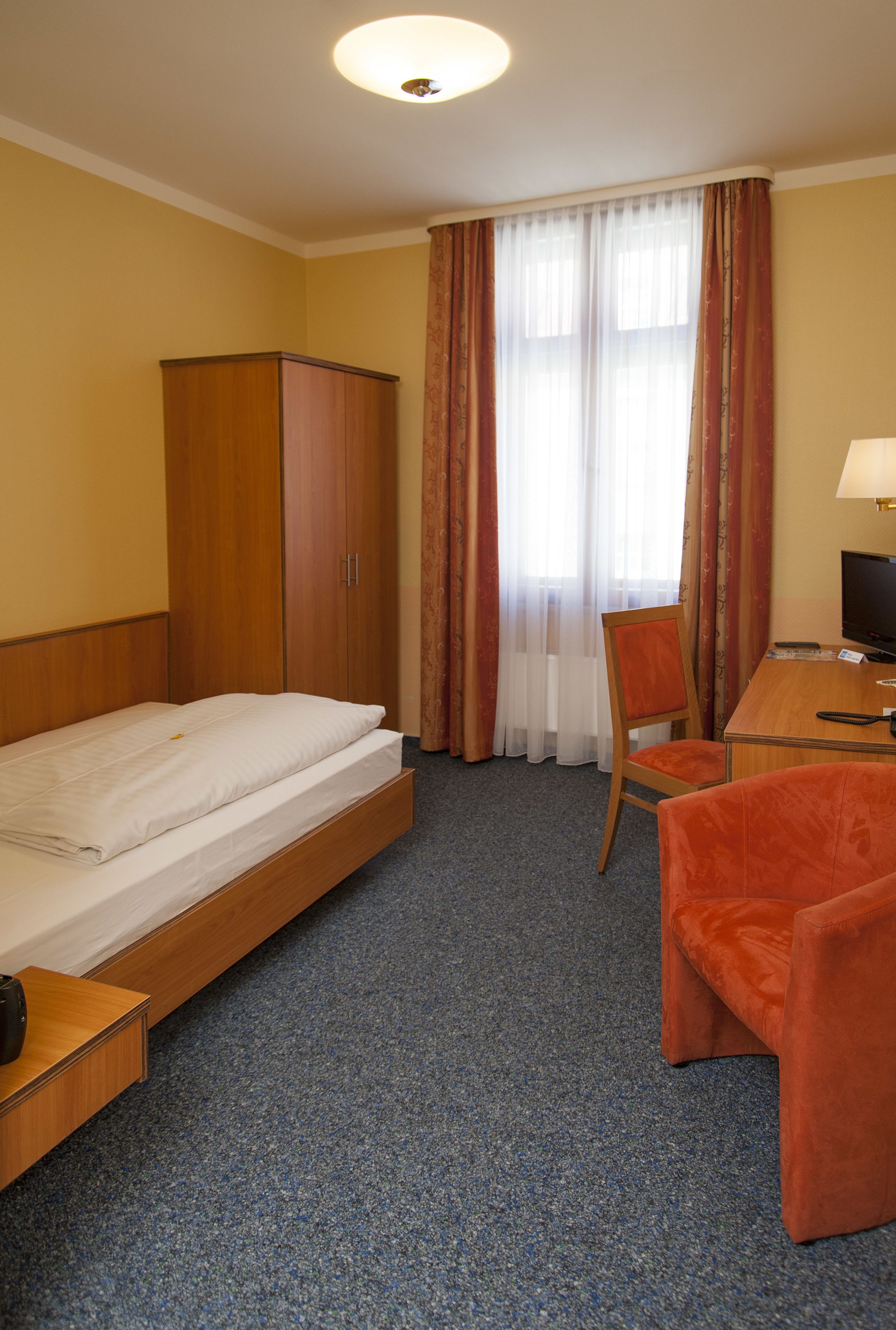 Quarto de hotel com cama, escrivaninha, cadeira, televisão e guarda-roupa.