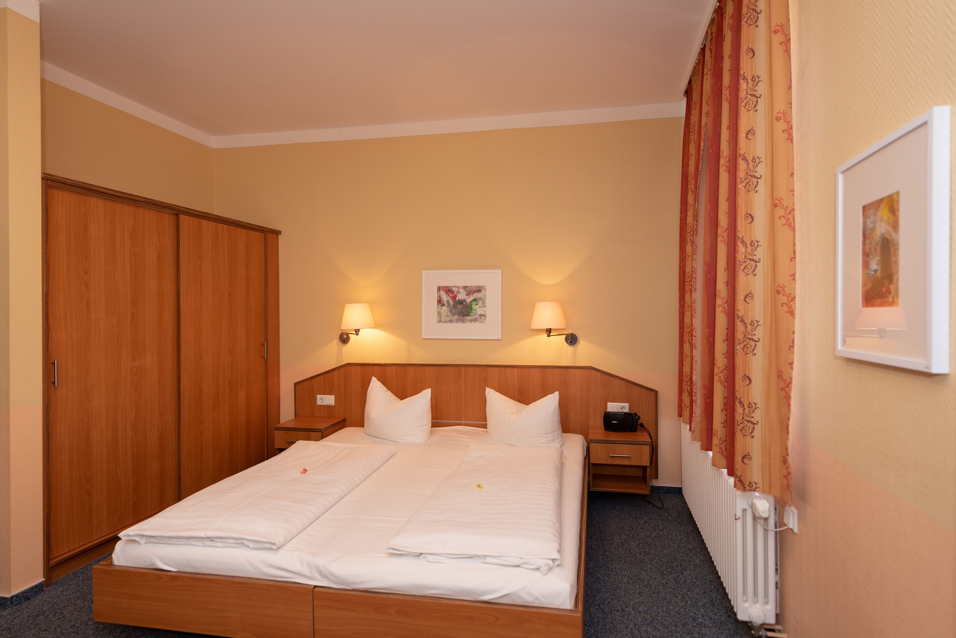 Habitación de hotel con cama doble, dos mesitas de noche, un armario de madera, un radiador y un cuadro en la pared.